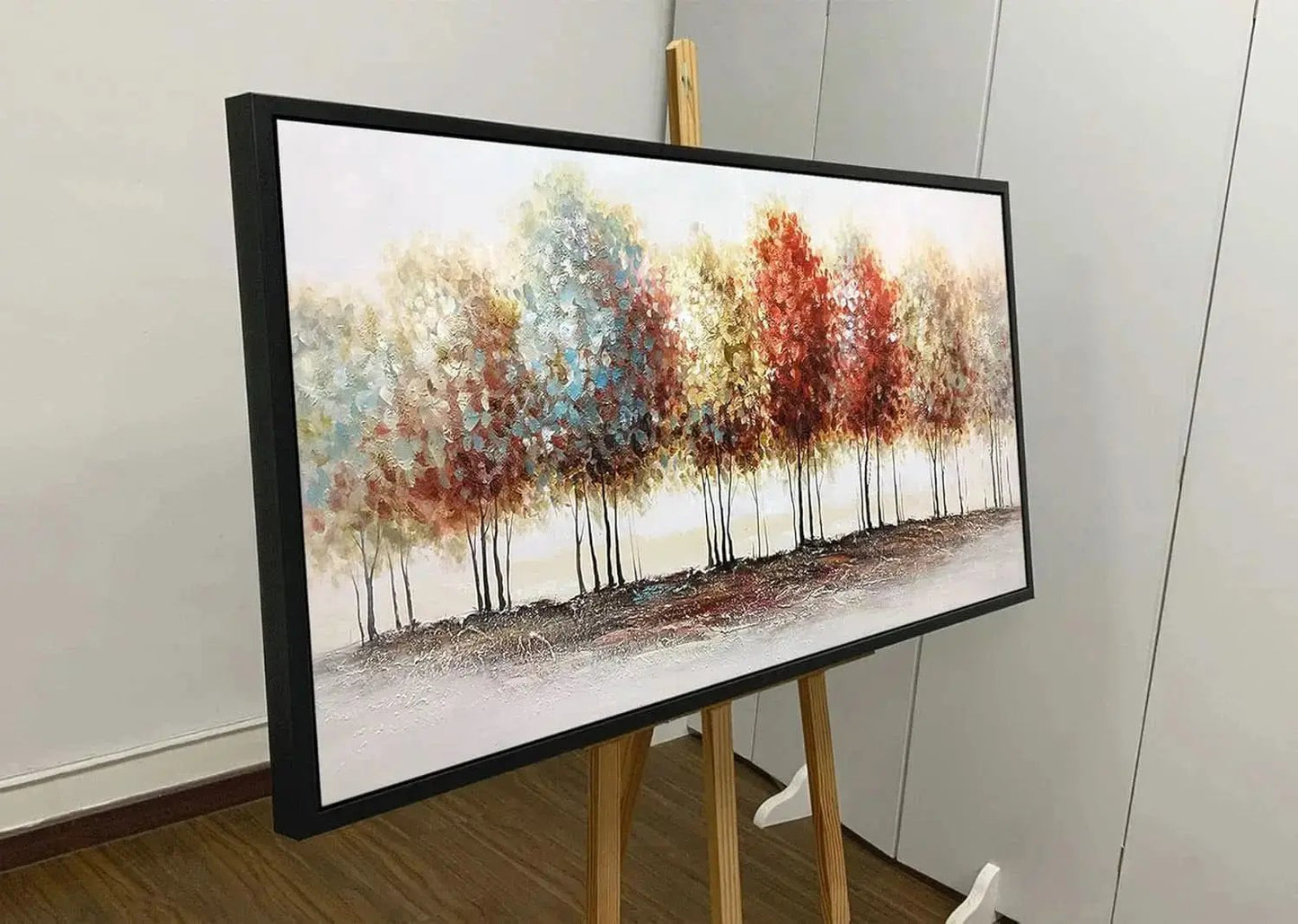 Framed artwork of trees with autumn colors on an easel against a white wall.