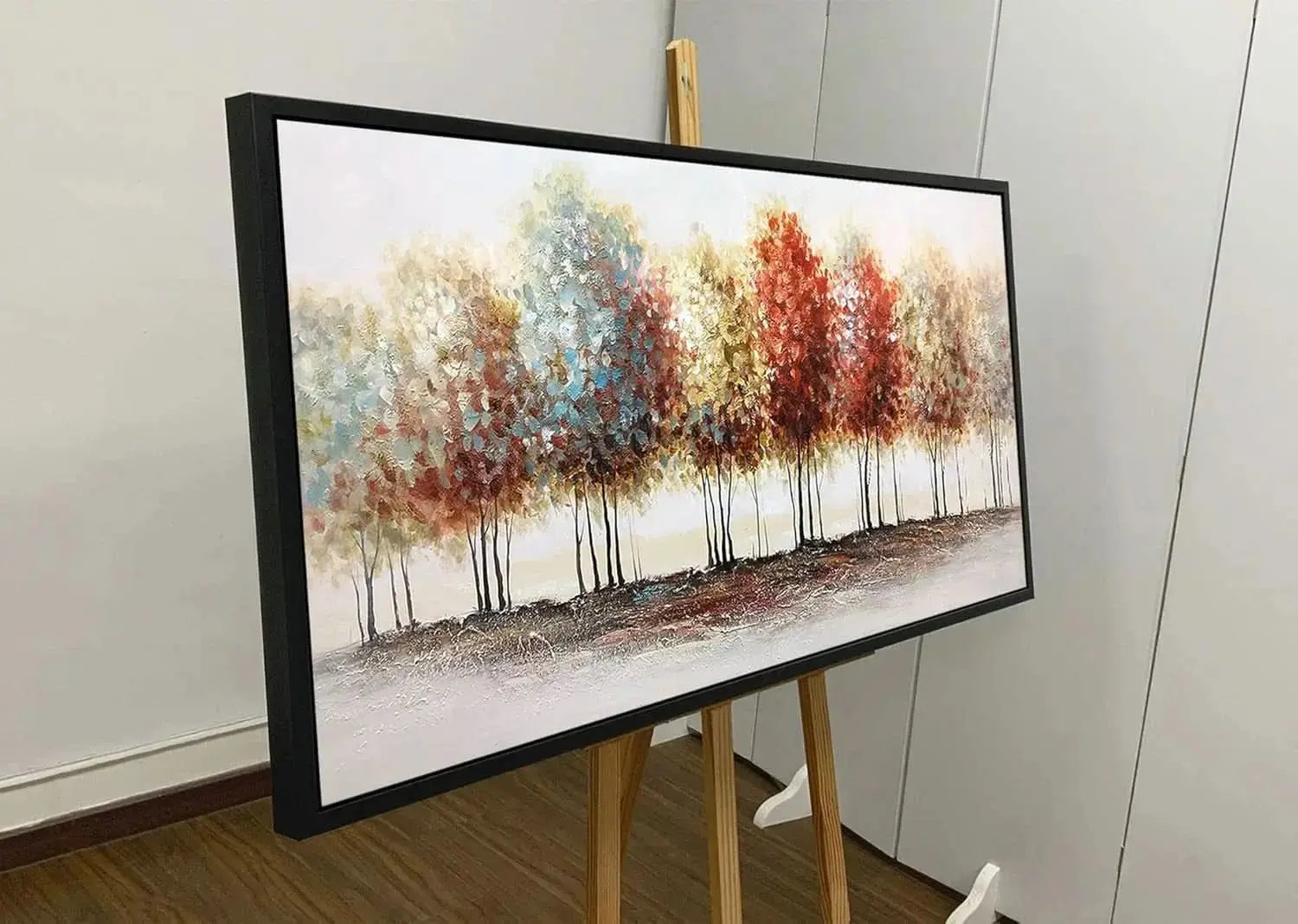 Framed artwork of trees with autumn colors on an easel against a white wall.