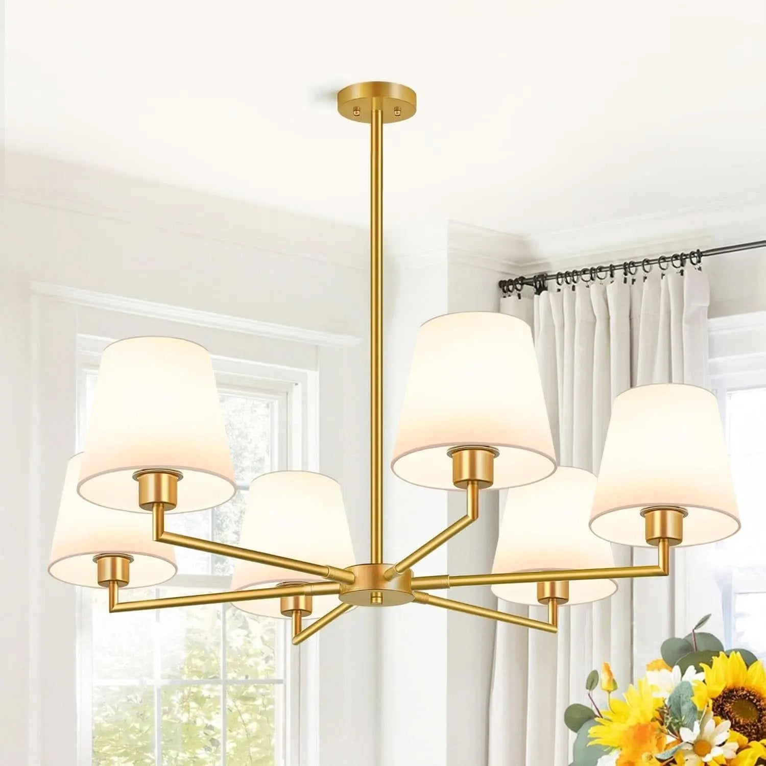 Gold chandelier with white lampshades in a room with curtains and flowers.