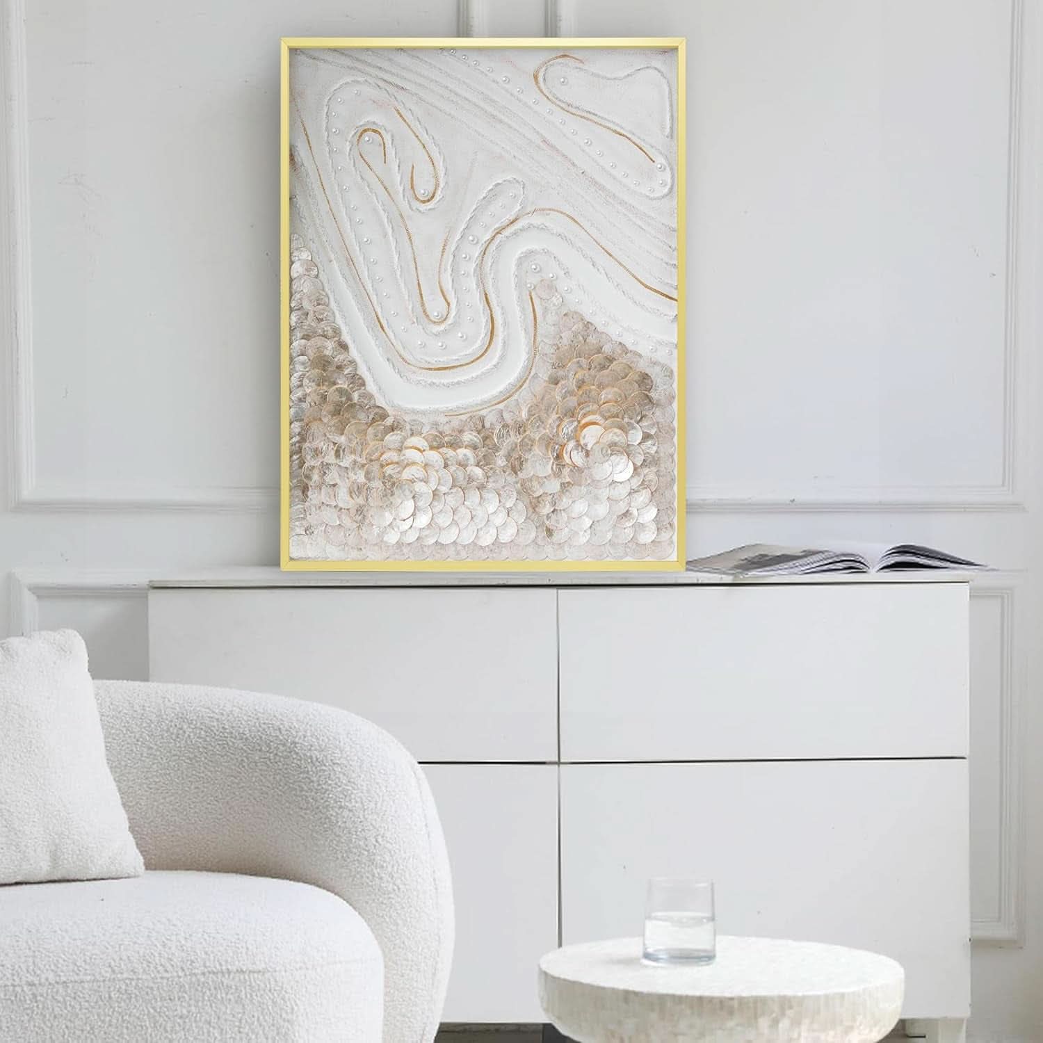 Abstract art piece in a gold frame on a wall above a white cabinet with a modern chair and small table.